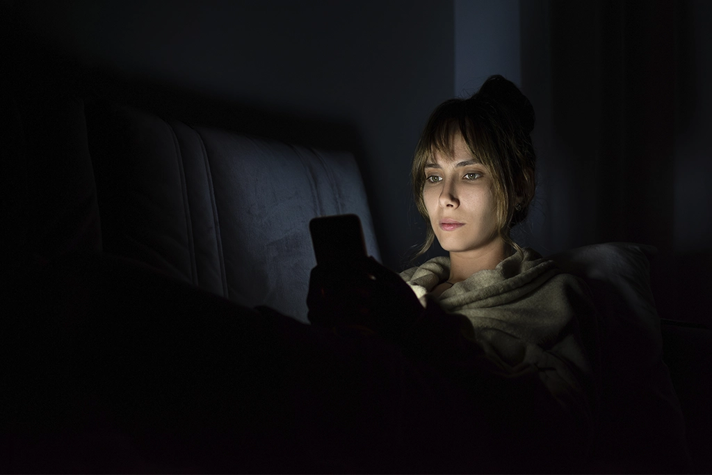 Mujer usando su celular en la oscuridad, ilustrando posibles daños por el uso excesivo del celular durante la noche.
