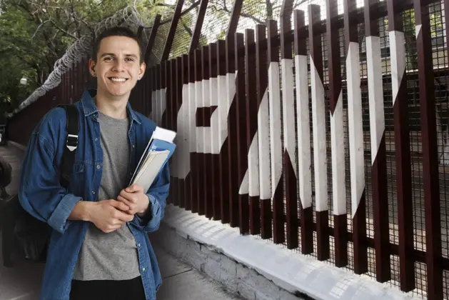Estudiante del IPN sosteniendo libros y sonriendo frente a una reja con el logo del Instituto Politécnico Nacional (IPN) para estudiar la nueva ingenieria