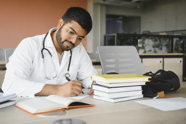 Joven estudiante de medicina revisando libros y un estetoscopio, enfrentando los retos de estudiar medicina en México: carga académica y financiera.