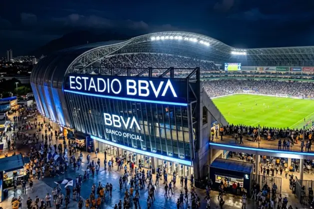 Estadio con nombre de banco iluminado durante un partido nocturno, mostrando la fachada del recinto deportivo con el logotipo de la institución financiera visible en primer plano.