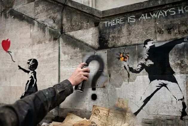 Mano de artista pintando un signo de interrogación en un mural, con documentos antiguos al pie, representando la búsqueda de cuál es la identidad de Banksy.
